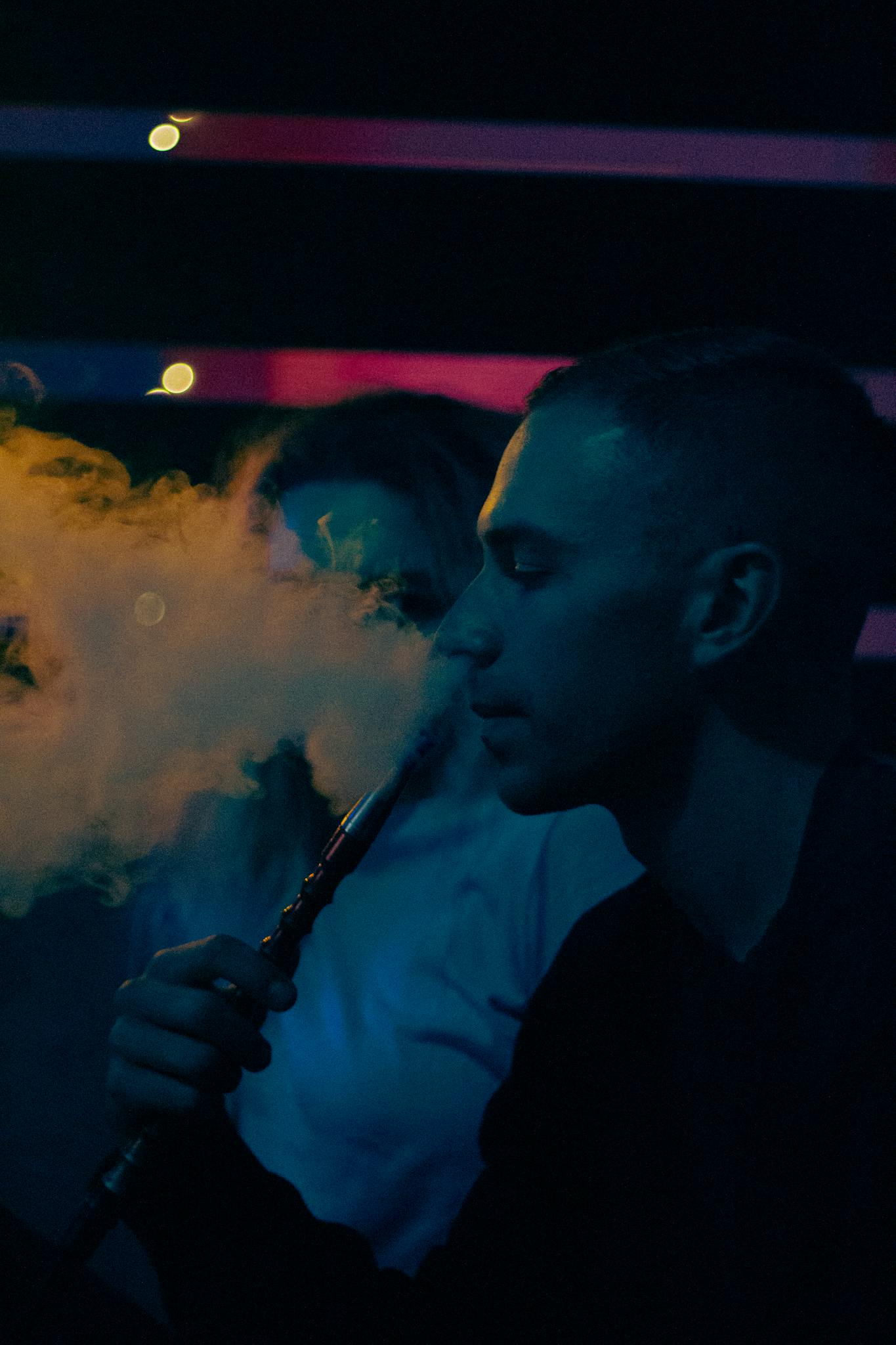 A young man smoking hookah in a moody, dark lounge with colored lighting.